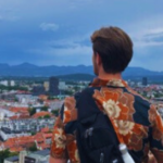 A student's back is in view as the student overlooks many buildings and a mountain range in the distance.