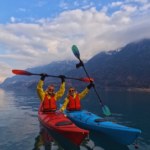 Two students in a red and blue kayak raise up their paddles in excitement as a photo is taken in still, clear water and vast clouds and mountains behind them