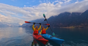 Two students in a red and blue kayak raise up their paddles in excitement as a photo is taken in still, clear water and vast clouds and mountains behind them