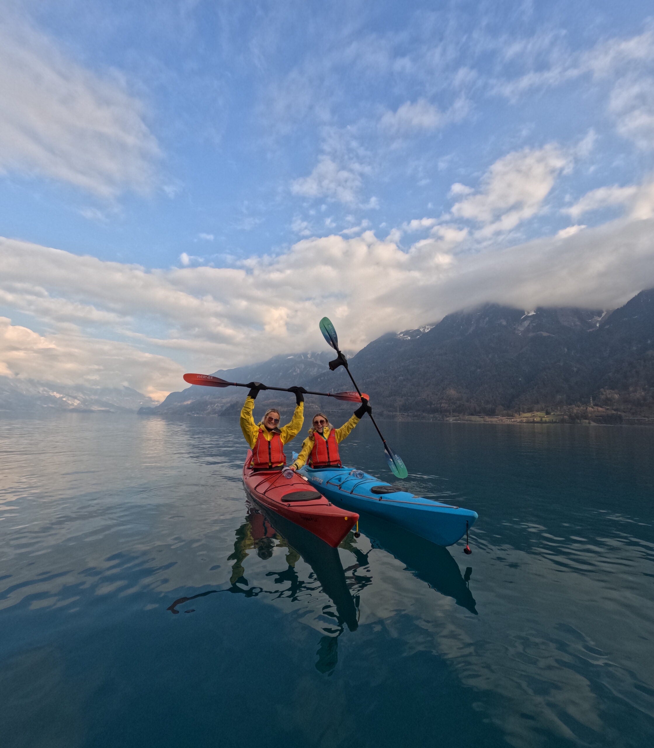Two students in a red and blue kayak raise up their paddles in excitement as a photo is taken in still, clear water and vast clouds and mountains behind them