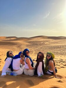 A group of female students wearing white and hijabs smile at the camera while sitting in the middle of a dessert