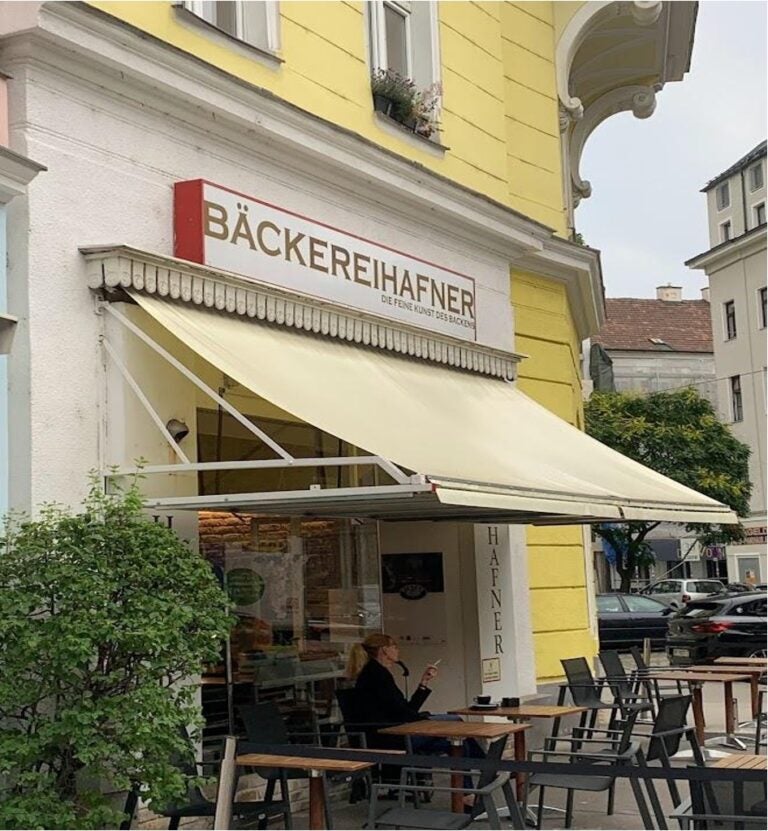 The outside of a shop has a yellow awning and a few wooden chairs and iron seats