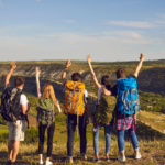 A small group of student travelers stand with their hands up in the air and travel backpacks around their back at the top of a valley or range