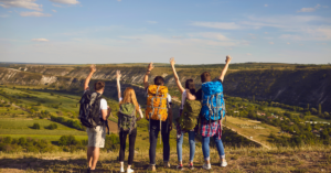 A small group of student travelers stand with their hands up in the air and travel backpacks around their back at the top of a valley or range