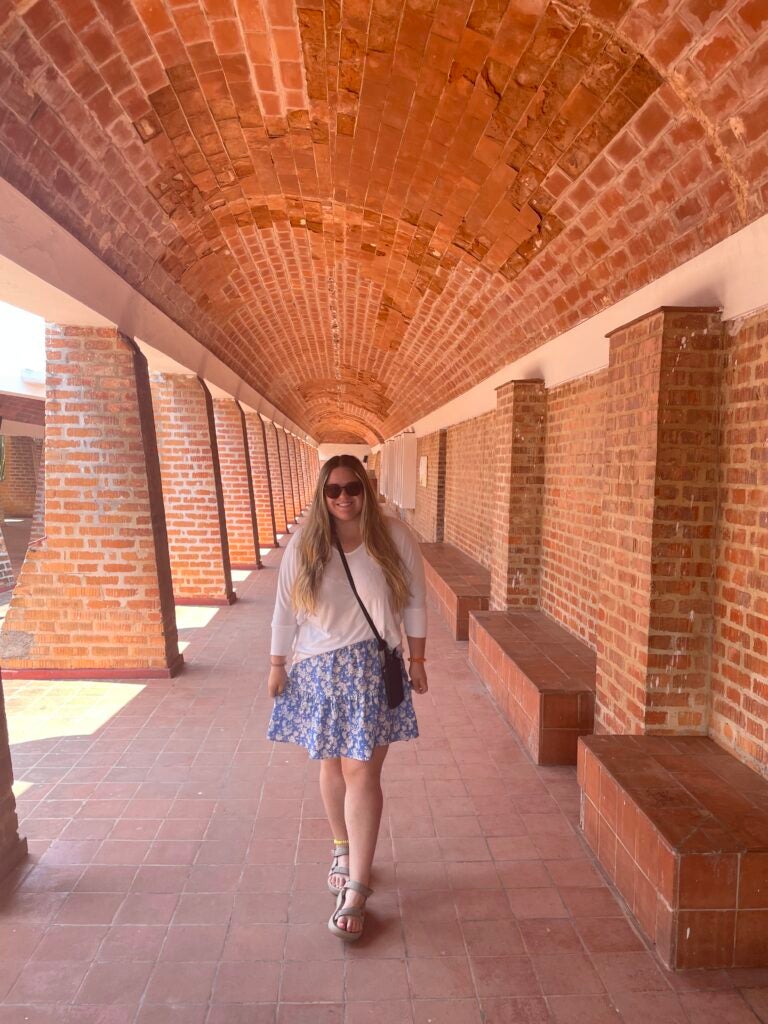 A student in a blue skirt, tennis shoes, and a white sweater smiles at a site in Cuba with several orange bricks displayed.