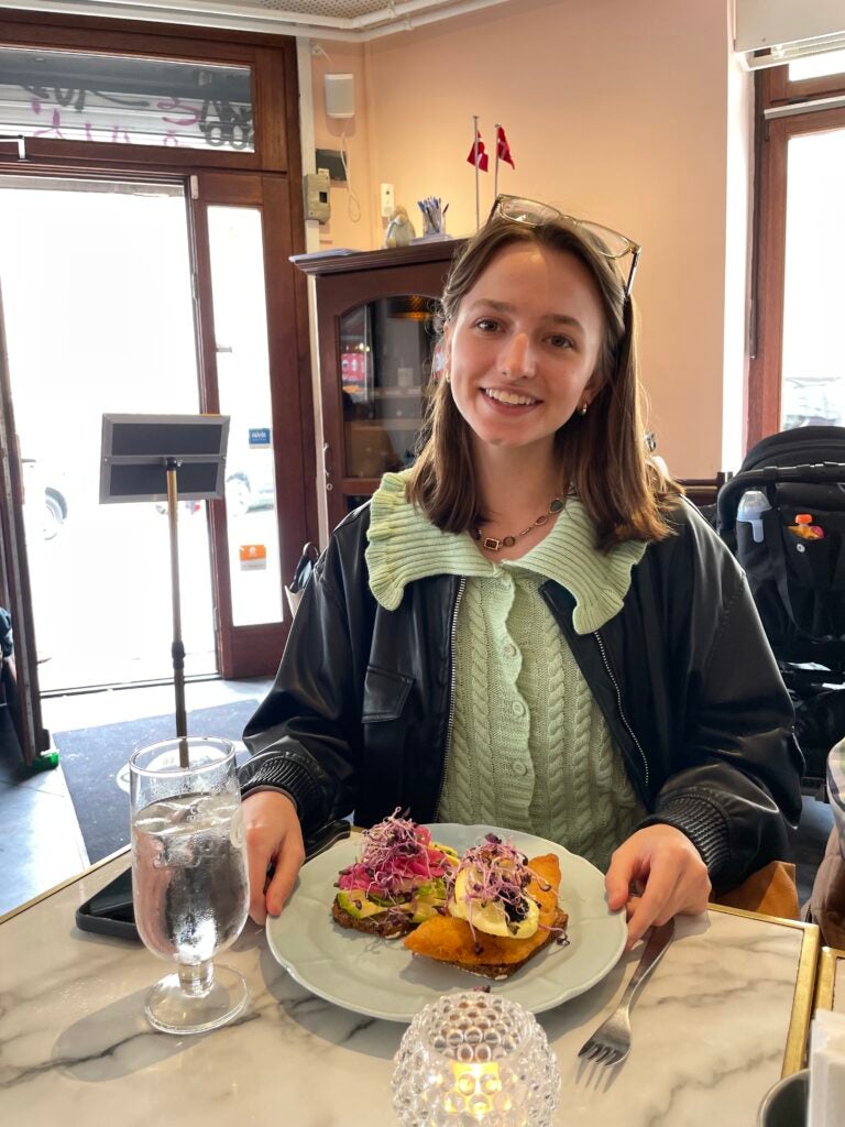 A student in a green top and black jacket smiles at the camera while eating at a restaurant.