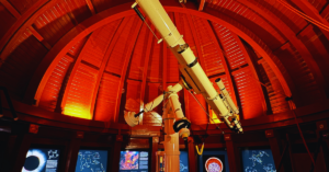 The inside roof of a planetarium is hues of red and maroon with a telescope in the center of the dome like room.