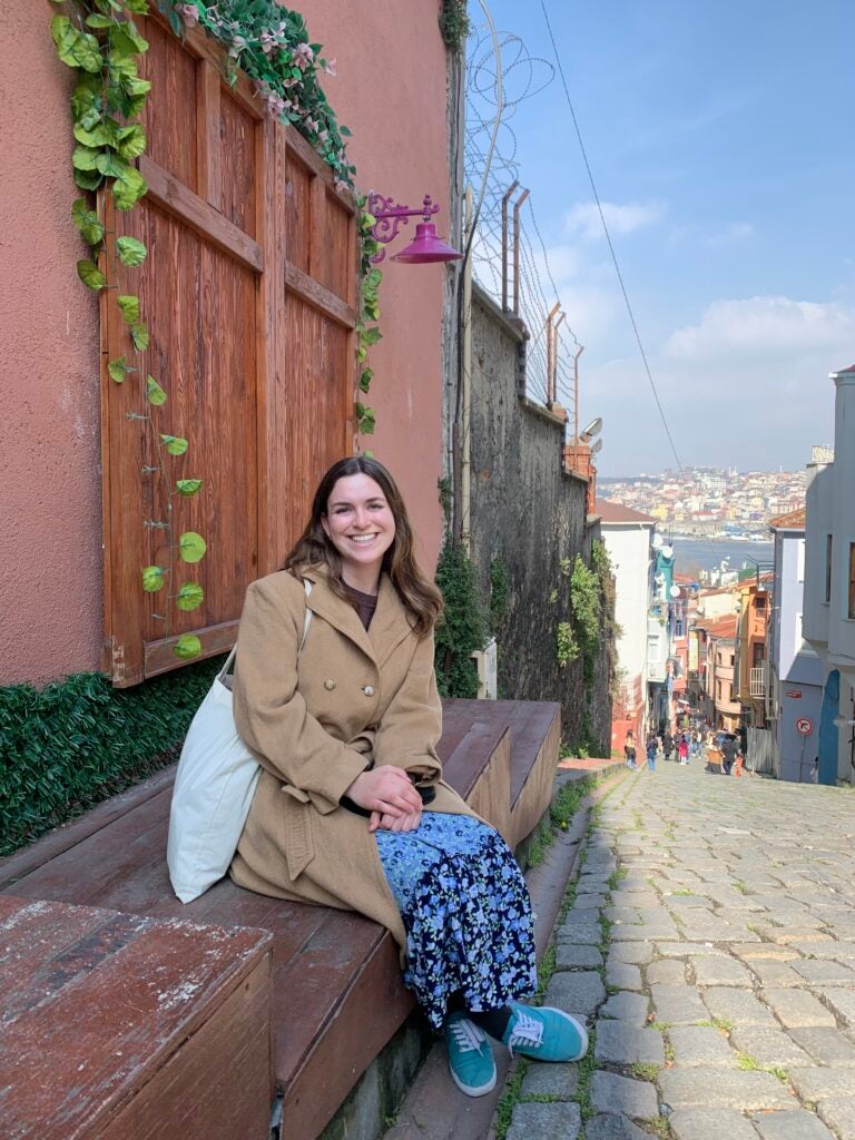 A student in a tan coat and blue pants smiles for a camera while sitting on a wooden bench on a small cobblestone road.