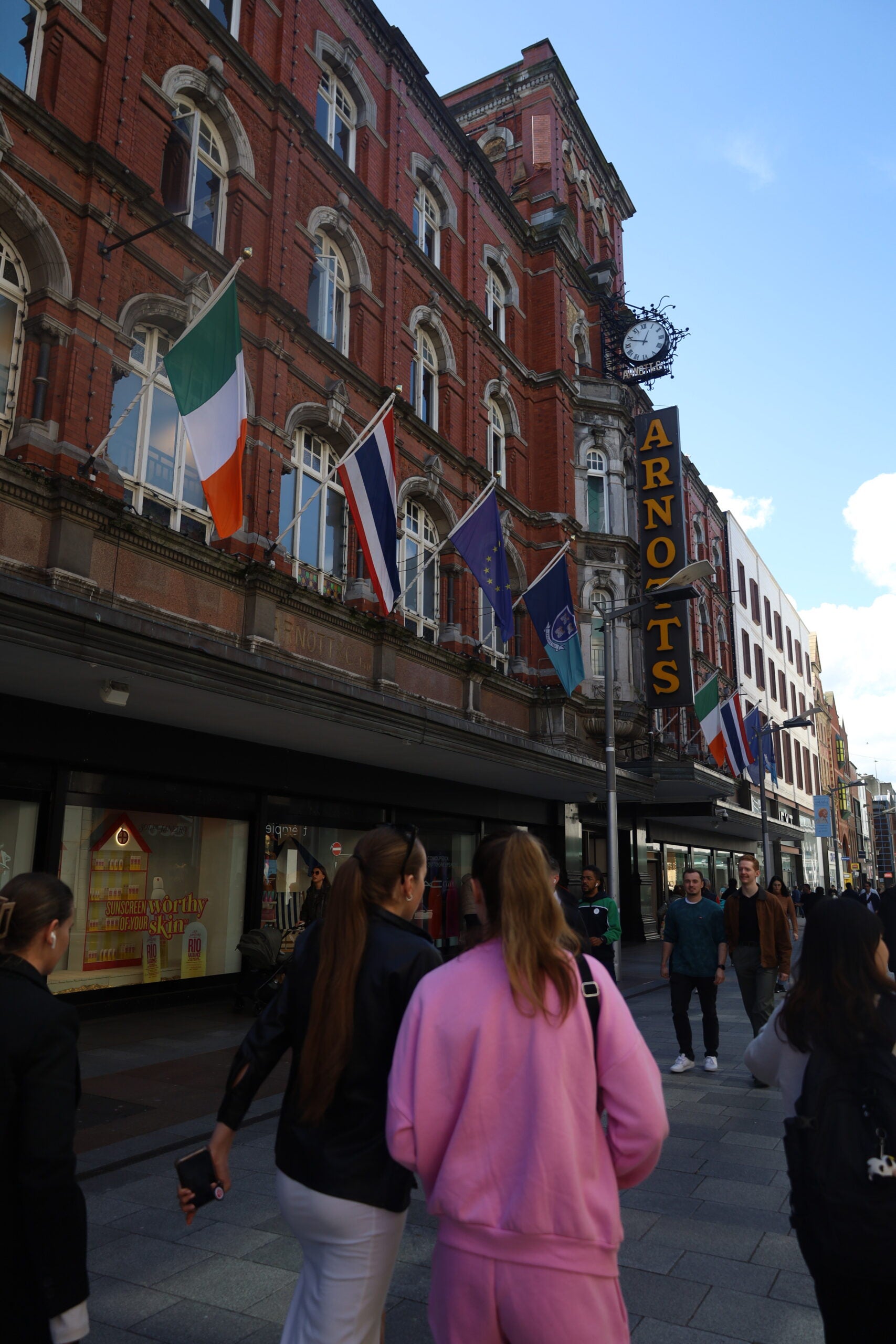 People walking along a city street with various international flags displayed on a building and a sign for "ARNOTTS" visible.