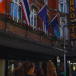 People walking along a city street with various international flags displayed on a building and a sign for "ARNOTTS" visible.