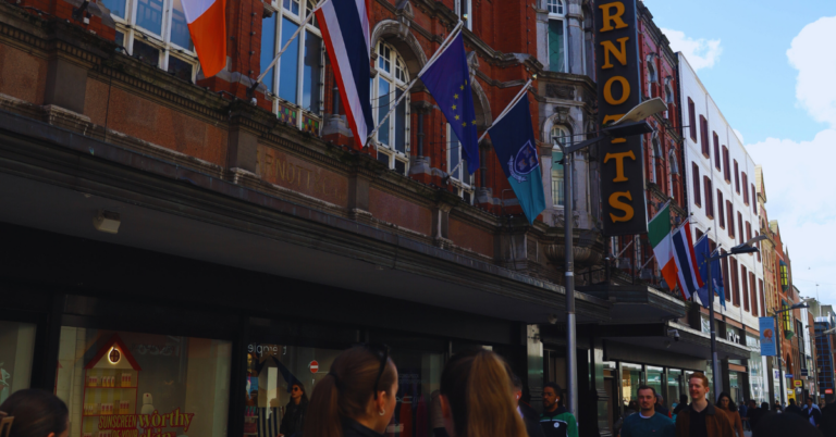 People walking along a city street with various international flags displayed on a building and a sign for "ARNOTTS" visible.