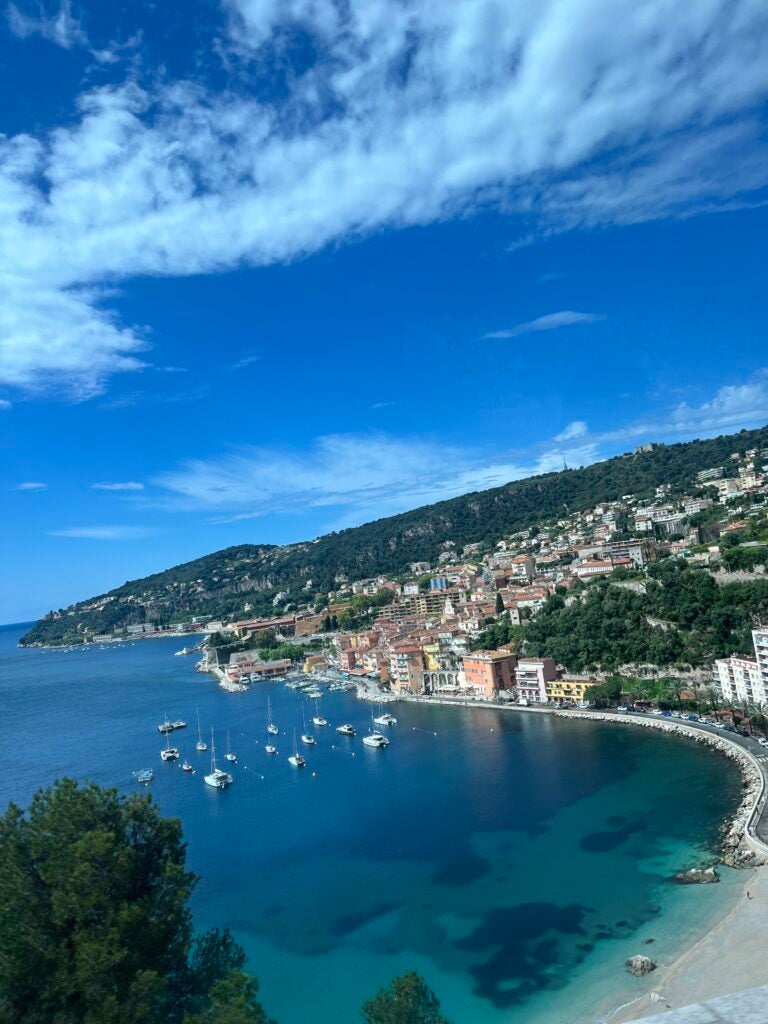 The cost of France has a few ships in the clear, blue water and many colorful buildings situated off the coast line.