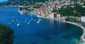 The cost of France has a few ships in the clear, blue water and many colorful buildings situated off the coast line.