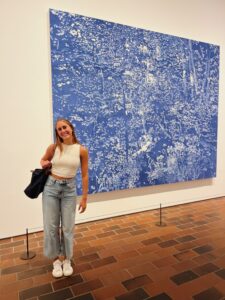 Student in white top and blue jeans and a black bag smiles at the camera with a large blue and white painting behind them.