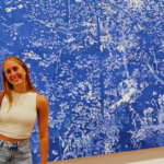 Student in white top and blue jeans and a black bag smiles at the camera with a large blue and white painting behind them.