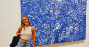 Student in white top and blue jeans and a black bag smiles at the camera with a large blue and white painting behind them.