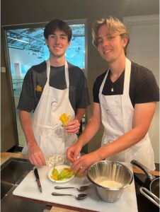 Two students smiling at the camera while making dinner wearing white aprons.