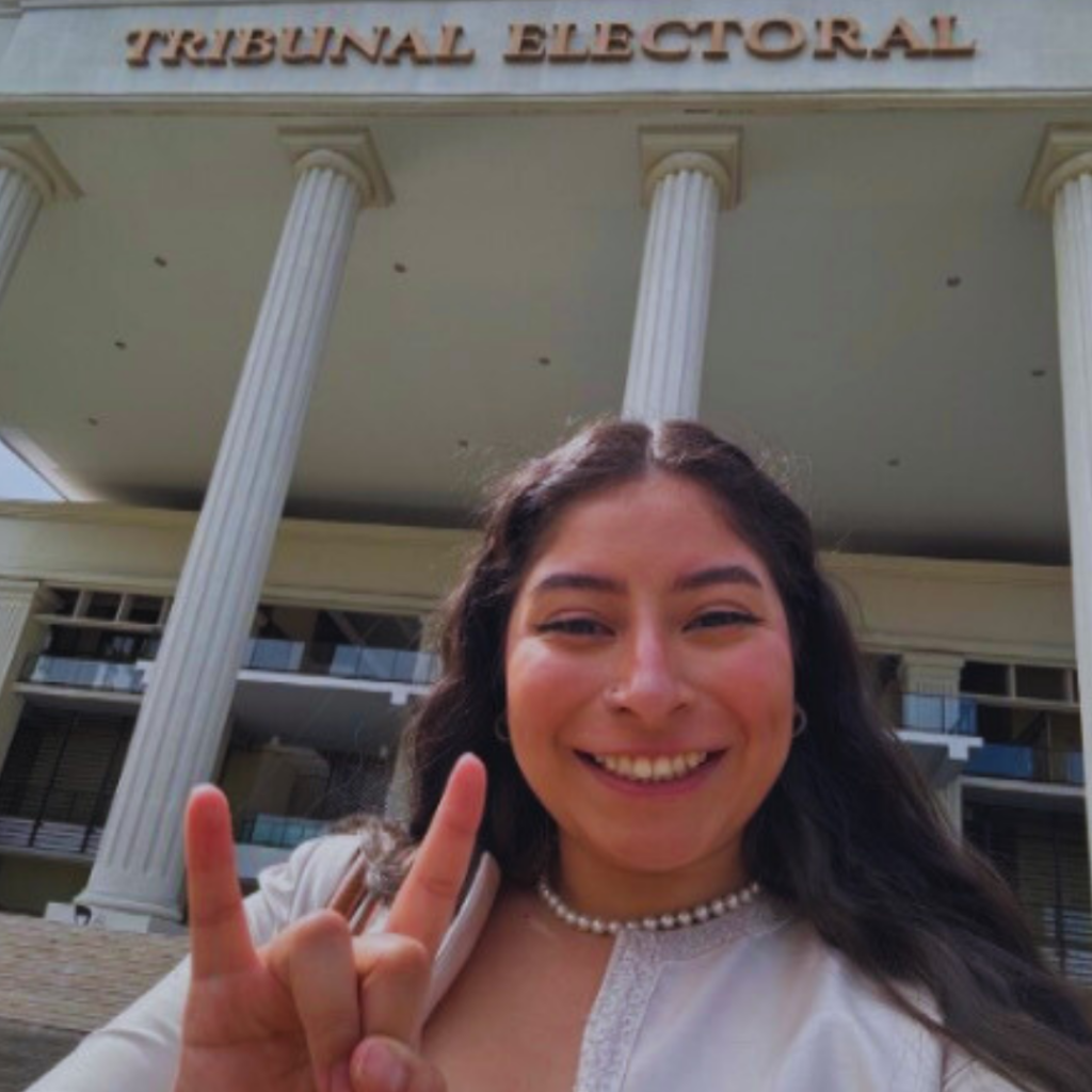 Student smiles in front of the Tribunal Electoral office holding up the longhorn hand sign.