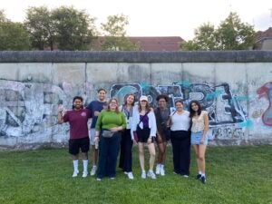 A group of students stand in front of the Berlin Wall where graffiti is written on the wall.