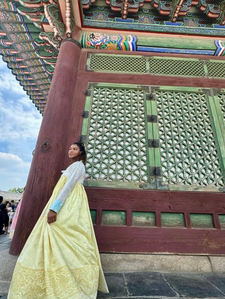 Student in traditional clothing of long colorful silk fabric stands outside of a traditional Korean building.