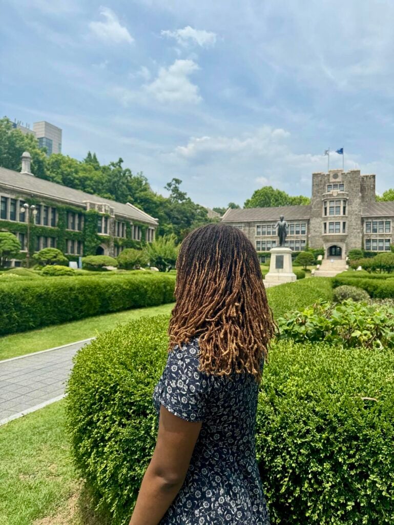 A students side profile is in view in front of a lush, green garden and large buildings