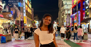 A student smiles directly at the camera in front of a bustling street in South Korea.