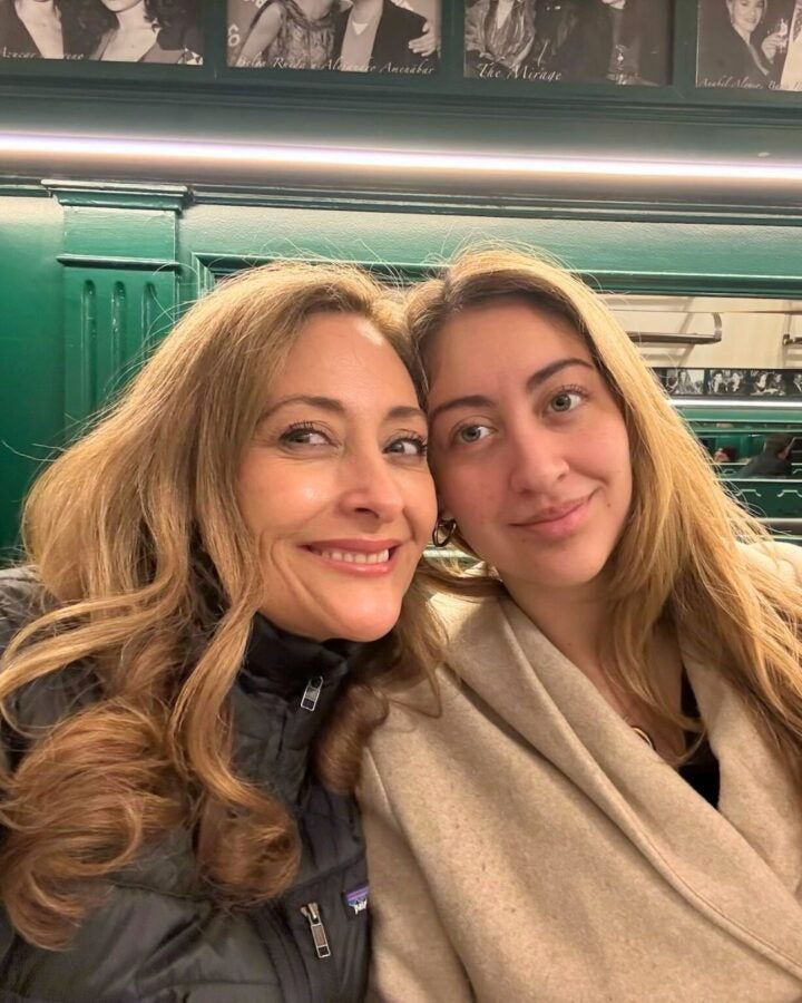 Isabella and her mother smiling at the camera, sitting closely on a green. Black-and-white photos are displayed on the walls behind them.