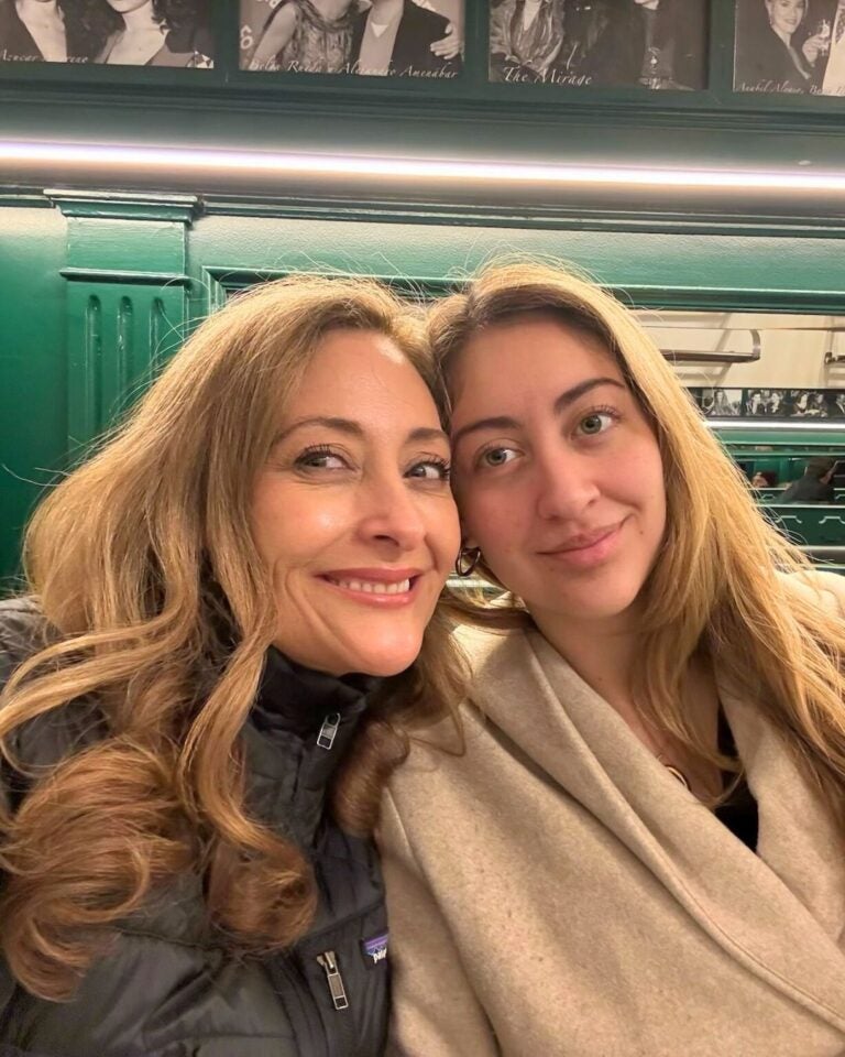 Isabella and her mother smiling at the camera, sitting closely on a green. Black-and-white photos are displayed on the walls behind them.