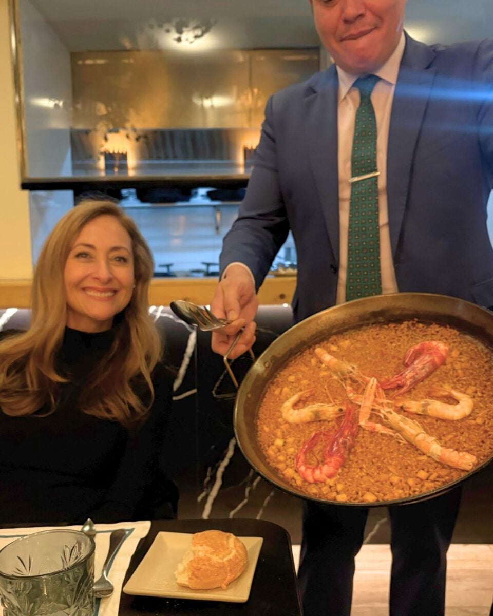 Isabella's mother at St. James Pontejos restaurant table as a server presents a pan of seafood paella. The dish is vibrant and garnished with large shrimp.