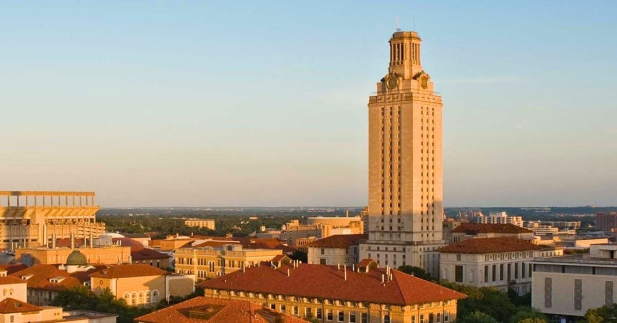 A tall, sunlit tower stands amidst a university campus with red-roofed buildings. The clear sky and warm lighting create a serene, academic atmosphere.