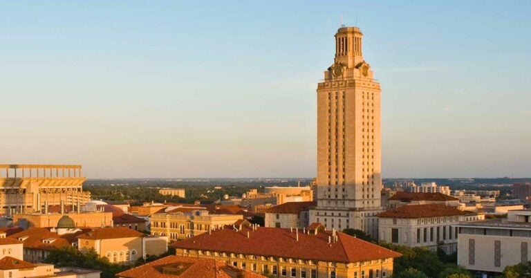 A tall, sunlit tower stands amidst a university campus with red-roofed buildings. The clear sky and warm lighting create a serene, academic atmosphere.