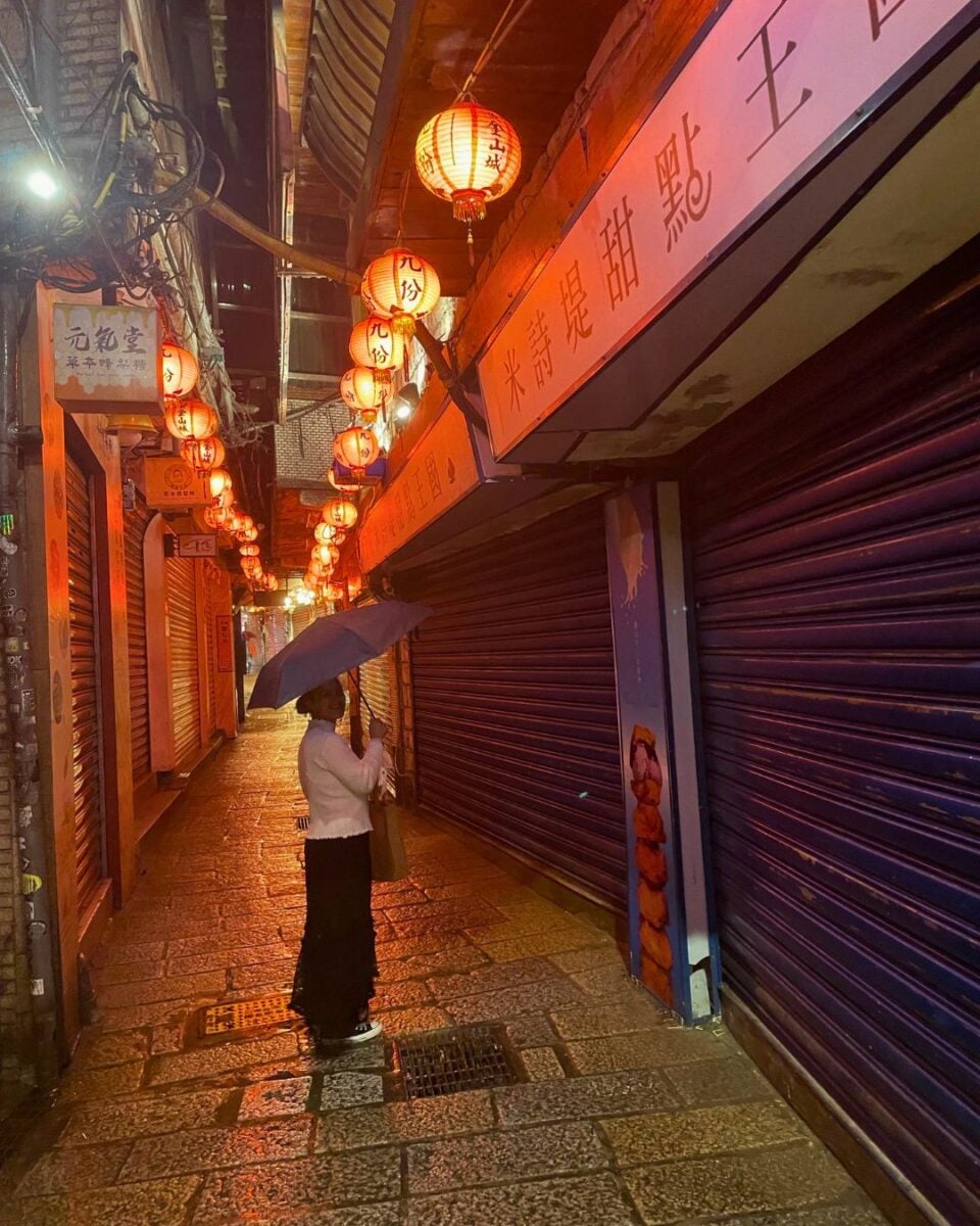 A person with an umbrella stands in a narrow alley lined with red lanterns, reflecting warm light on wet cobblestones. The street feels quiet and atmospheric.