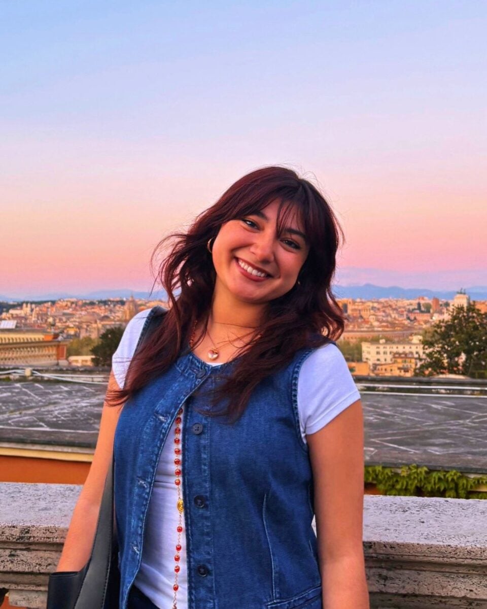 Smiling woman in a denim vest stands against a vibrant sunset backdrop, cityscape below. The scene exudes warmth and joy, highlighting her cheerful expression.