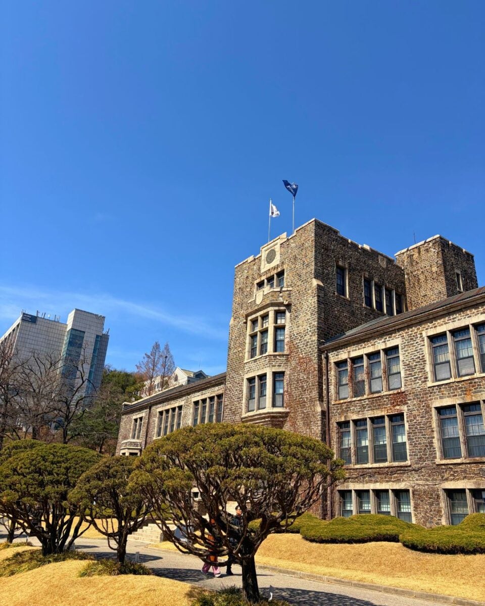 A historic stone building with flags atop stands under a clear blue sky. Trimmed trees and dry grass in the foreground suggest a peaceful, sunny day.