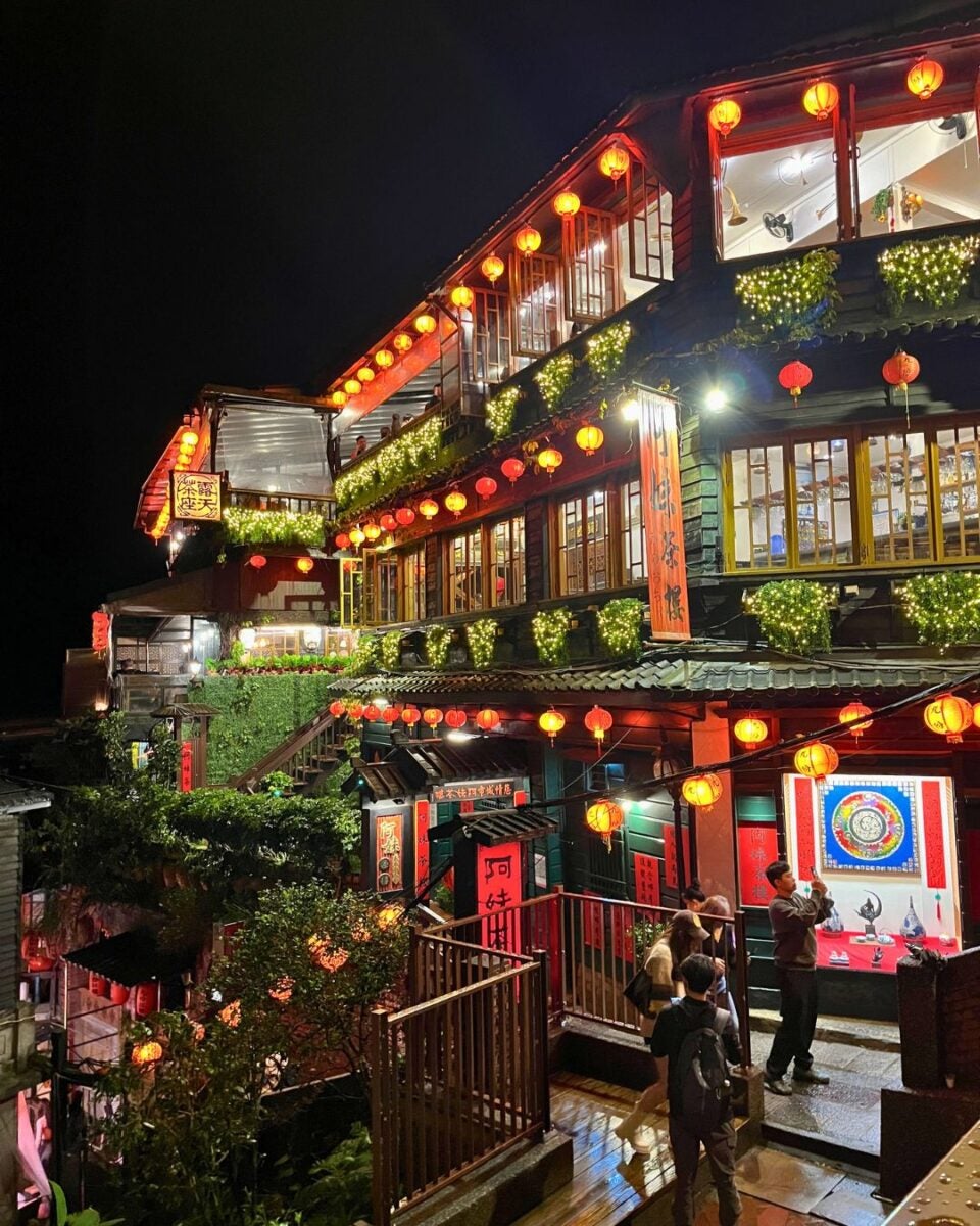 A beautifully lit, traditional tea house at night featuring red lanterns and lush greenery. Visitors admire the building's vibrant, inviting atmosphere.