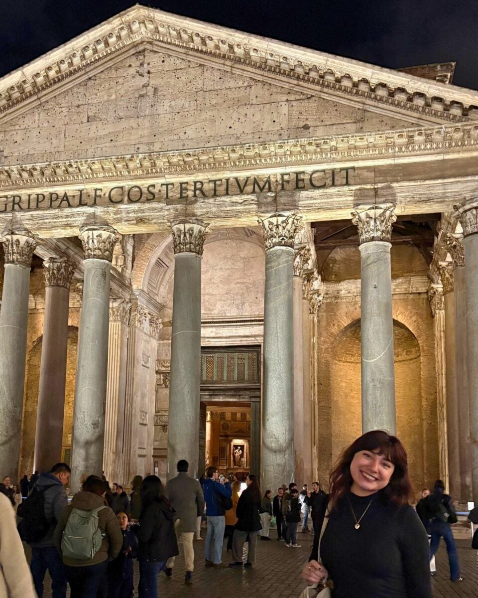 Smiling person in front of the Pantheon in Rome at night. The ancient building's illuminated columns and inscriptions are visible, with a lively crowd around.