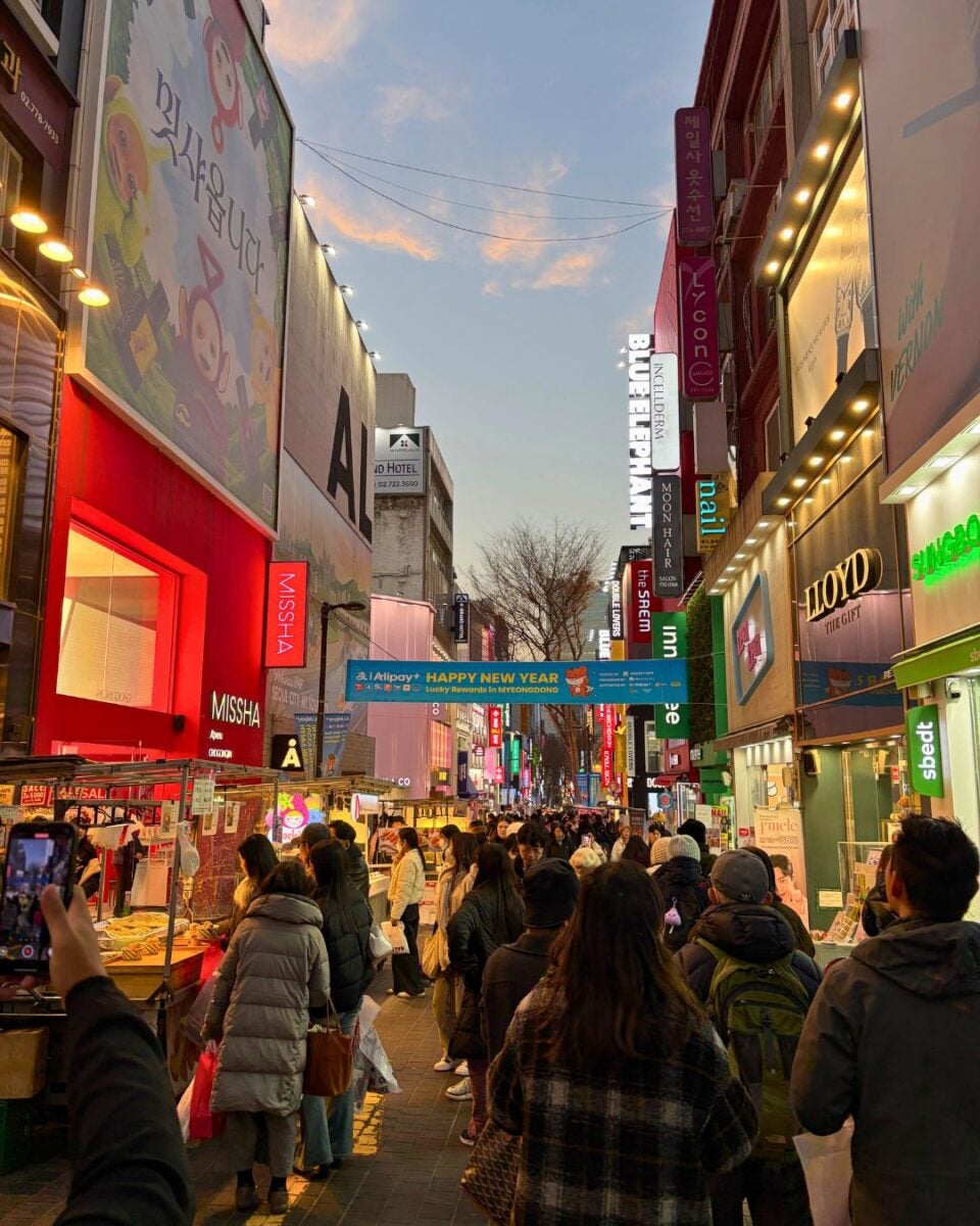 Bustling street scene in Myeongdong, Seoul at dusk, crowded with people shopping. Colorful neon signs illuminate food stalls and retail stores, creating a lively atmosphere.