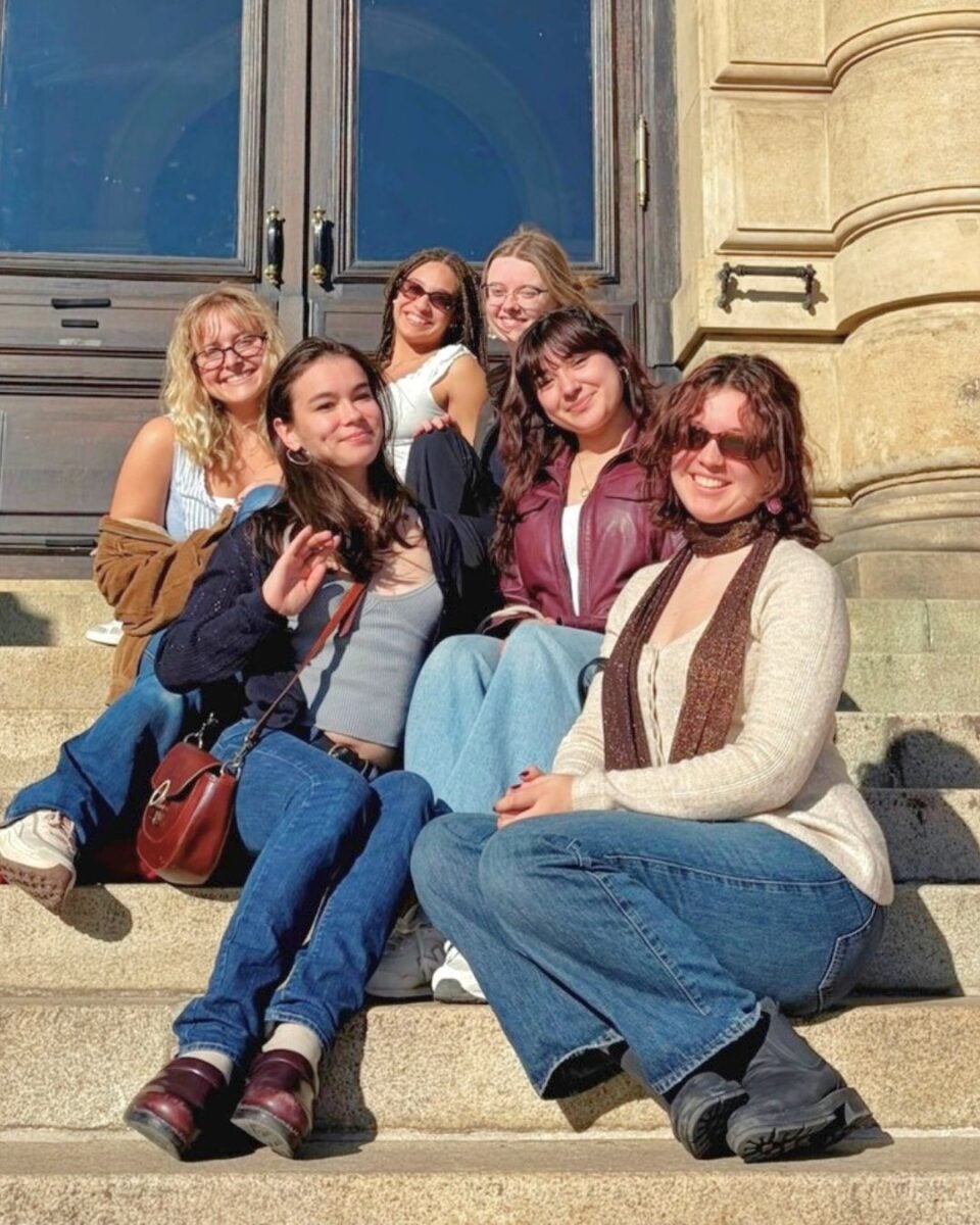 A group of six young women sits on stone steps outside a building.
