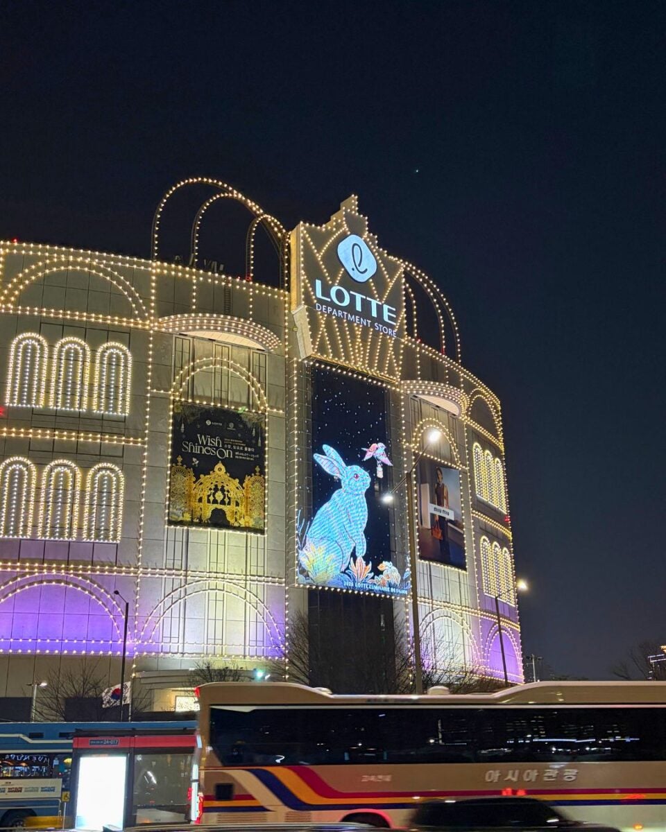 Illuminated Lotte Department Store at night with festive lights, featuring a large glowing rabbit display and vibrant city buses passing by.