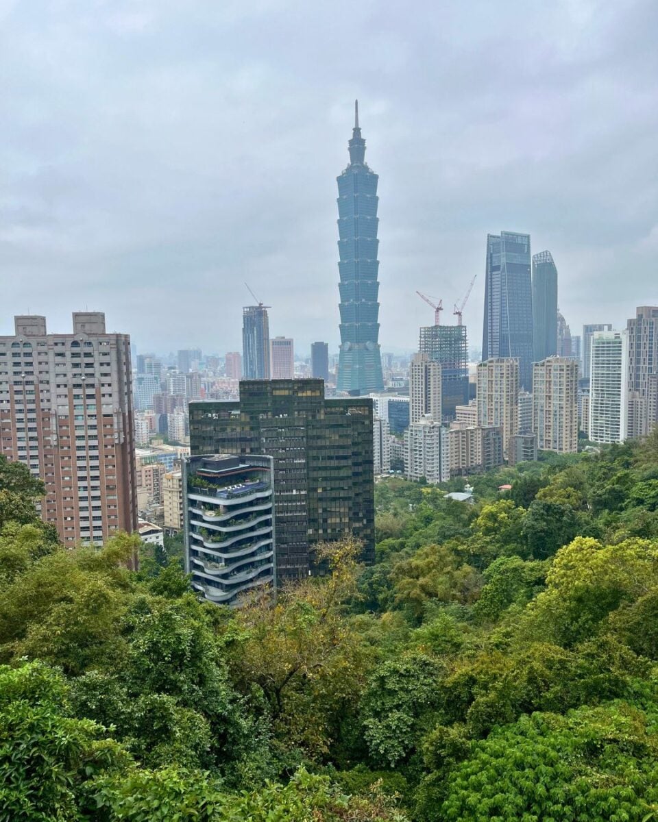Skyline of a city on a cloudy day, dominated by a distinctive tall skyscraper. Lush green trees are in the foreground, adding contrast. Urban and natural blend conveying a calm, balanced atmosphere.