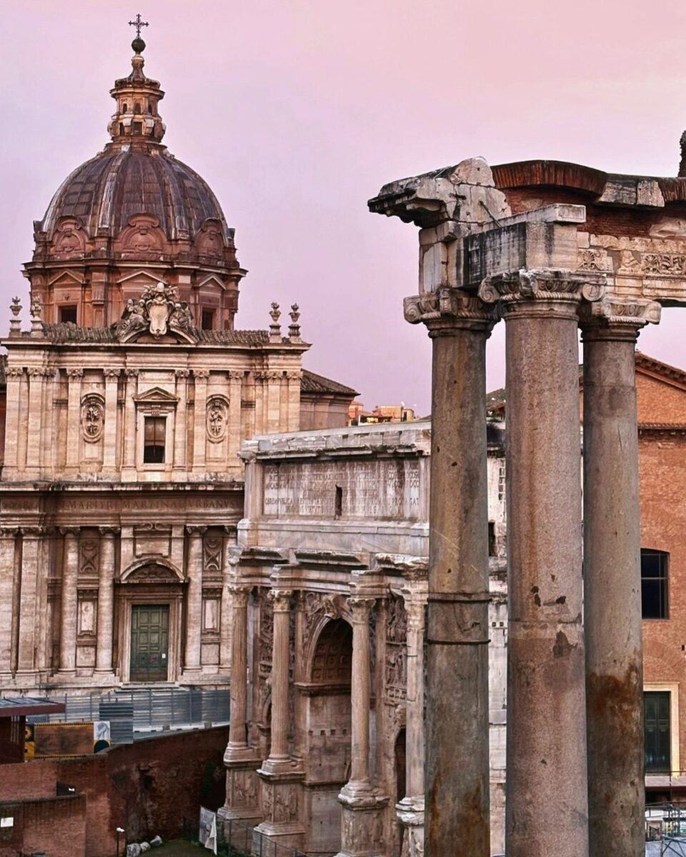 Dome of a baroque church with ornate columns in the foreground against a pastel pink sky, evoking a sense of historic elegance and timeless beauty.