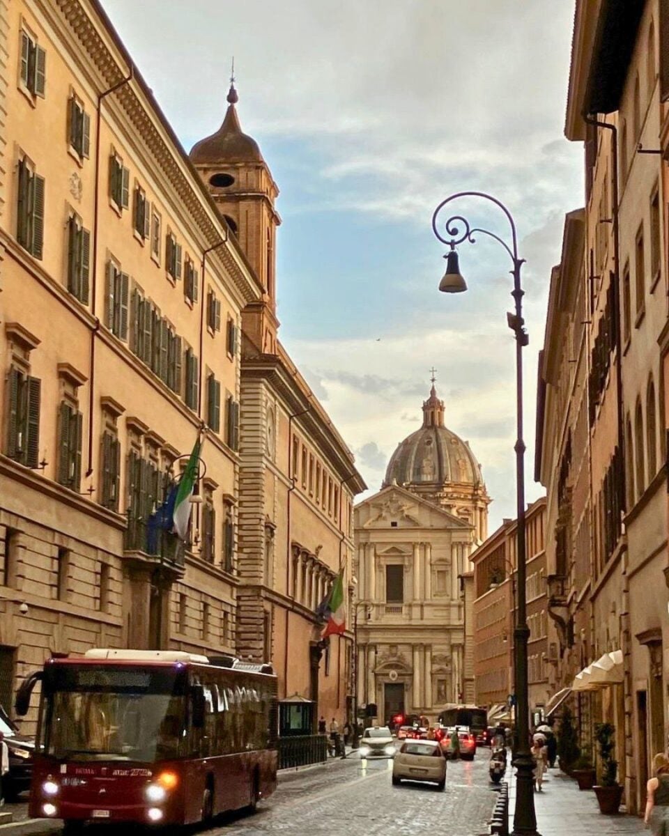 Street scene in Rome with a red bus and cars on a cobblestone road. Flanked by historic buildings, it leads to a domed church under a cloudy sky.