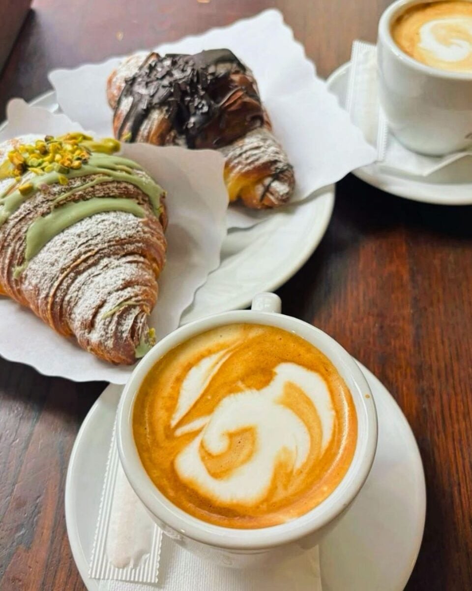 Two croissants, one with pistachio and the other with chocolate, are on a table with two cappuccinos featuring swirl patterns, creating a cozy breakfast scene.