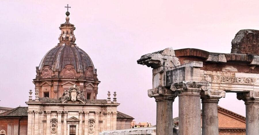 Dome of a baroque church with ornate columns in the foreground against a pastel pink sky, evoking a sense of historic elegance and timeless beauty.