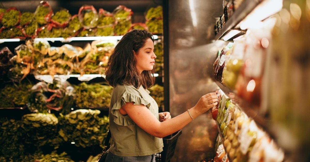 A woman with dark hair selects items from a well-lit grocery aisle, surrounded by fresh produce and packaged goods. The scene conveys calm and focus.
