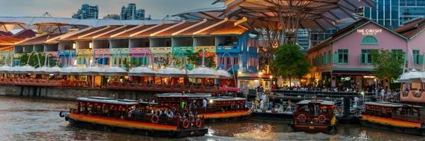 A lively riverside with boats docked, colorful buildings, and an open-air restaurant at dusk.