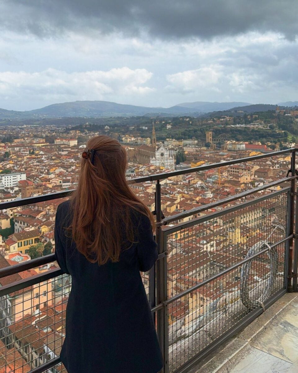 Person overlooking a cityscape with historic buildings and a cathedral under a cloudy sky.