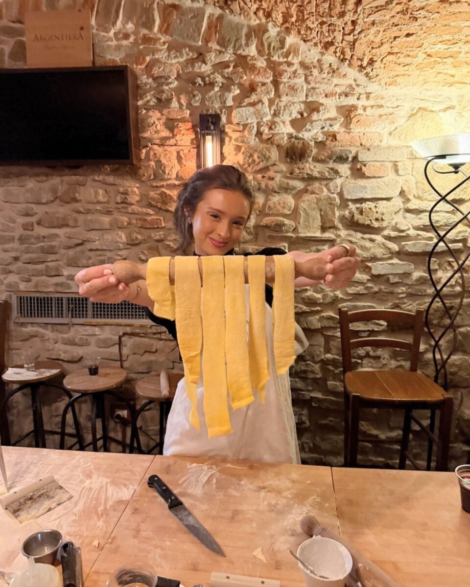 A woman holding strips of fresh pasta on a rolling pin in a rustic kitchen with stone walls.