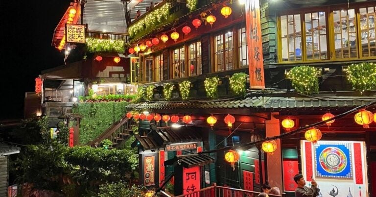 Amei Tea House is beautifully lit at night featuring red lanterns and lush greenery. Visitors admire the multi-story building's vibrant, warm, and inviting atmosphere.