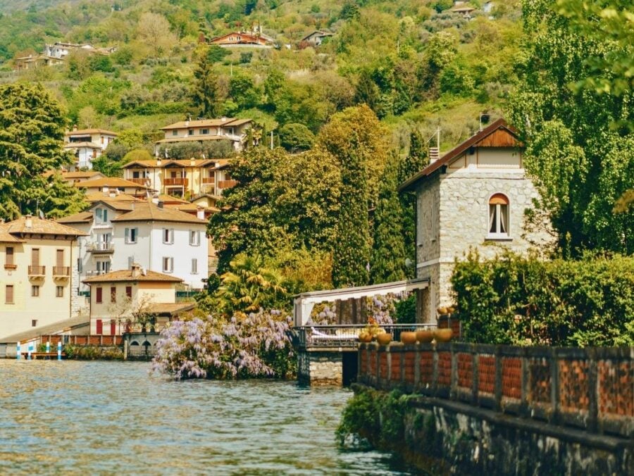 Charming lakeside European village with colorful houses surrounded by lush greenery. A peaceful narrow waterfront path leads past vibrant plants and wisteria.